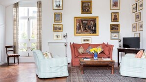 Un salon avec un canapé rouge, deux fauteuils, une table basse et un téléviseur. Les murs sont ornés de tableaux encadrés et un lustre est suspendu au plafond.
