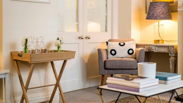 Une pièce chaleureuse avec un fauteuil gris, une table d’appoint en bois sur laquelle sont posés des verres, une lampe et une bibliothèque.