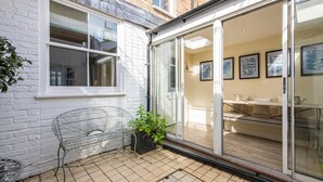 Un espace patio agrémenté d’un mur en briques blanches, d’une chaise en métal, d’une plante en pot et d’une porte-fenêtre coulissante donnant sur un espace intérieur avec un canapé et des œuvres d’art encadrées.