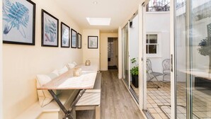 Un couloir étroit avec une longue table-banc en bois, des reproductions botaniques encadrées accrochées au mur et une porte vitrée donnant sur un patio extérieur.