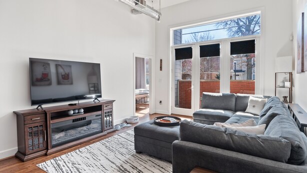 A modern living room with a large flat-screen TV, dark wood furniture, a grey sofa, and a patterned rug. - Spacious Condo in Center of NULU with Free, Gated, Secure Parking and Entrance (Louisville)
