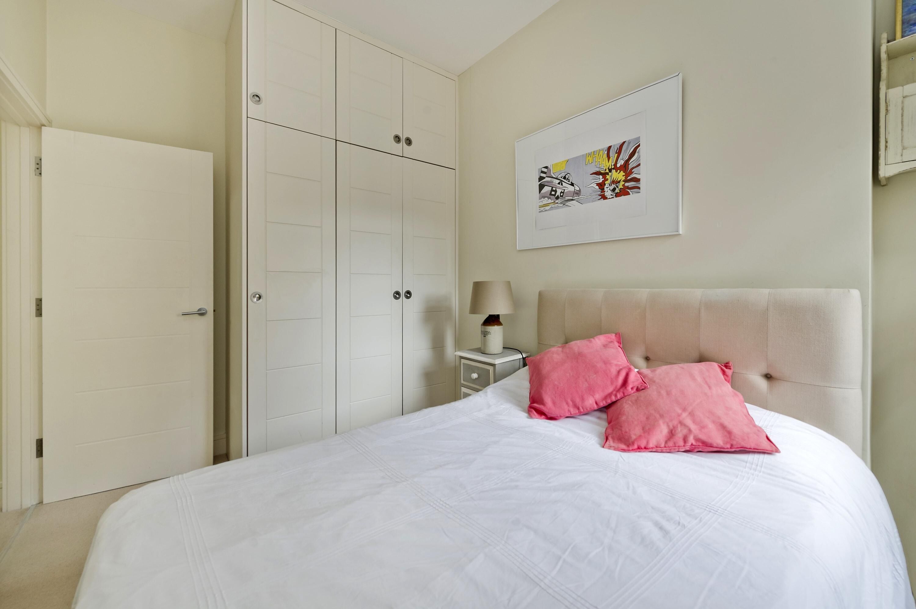 A bedroom with a bed, white walls, a wardrobe, a bedside table with a lamp, and a framed picture on the wall.