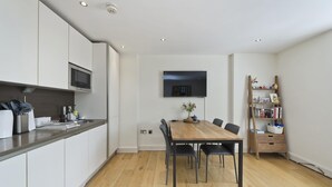 A modern kitchen with white cabinets, a wooden dining table, and a flat-screen TV mounted on the wall.