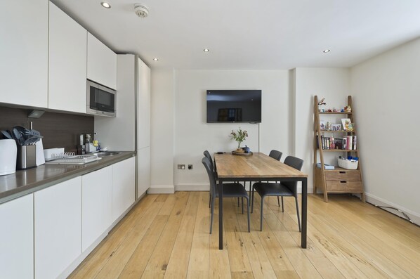 A modern kitchen with white cabinets, a wooden dining table, and a flat-screen TV mounted on the wall.