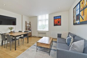 A modern living room with a dining area, a grey sofa, a wooden coffee table, and a large window with blinds.
