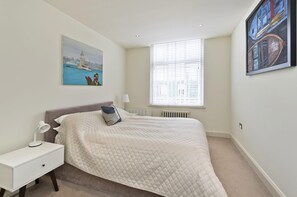 A bedroom with a bed, a nightstand, a lamp, a window with blinds, and two paintings on the walls.