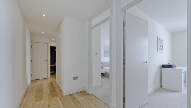 A hallway with wooden flooring leading to a bedroom with a bed and a desk.