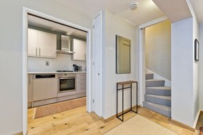 Una cocina moderna con armarios blancos, horno empotrado y cafetera. Hay una pequeña mesa y una alfombra en el suelo. Se ve una escalera a la derecha.