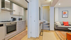 A modern kitchen with white cabinets, a stove, and an oven. There is a door leading to a staircase and a living area with a sofa and a painting on the wall. - Sweet Marble Arch 2 Bedroom Mews House (London)