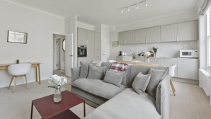 Uma sala moderna com um sofá cinzento, uma mesa pequena de madeira e uma mesa de centro vermelha. Há uma área de cozinha com armários brancos e um micro-ondas.