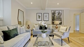 A modern living room with a grey sofa, a glass coffee table with a white vase, and a grey armchair. There are framed botanical prints on the wall and a large window with a view of trees. - Huge Kensington Garden Flat (London)