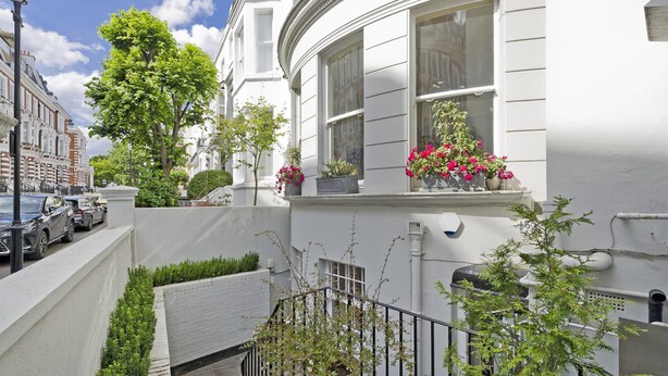 A white townhouse with a curved facade, large windows, and a small balcony with potted plants and flowers. - Huge Kensington Garden Flat (London)