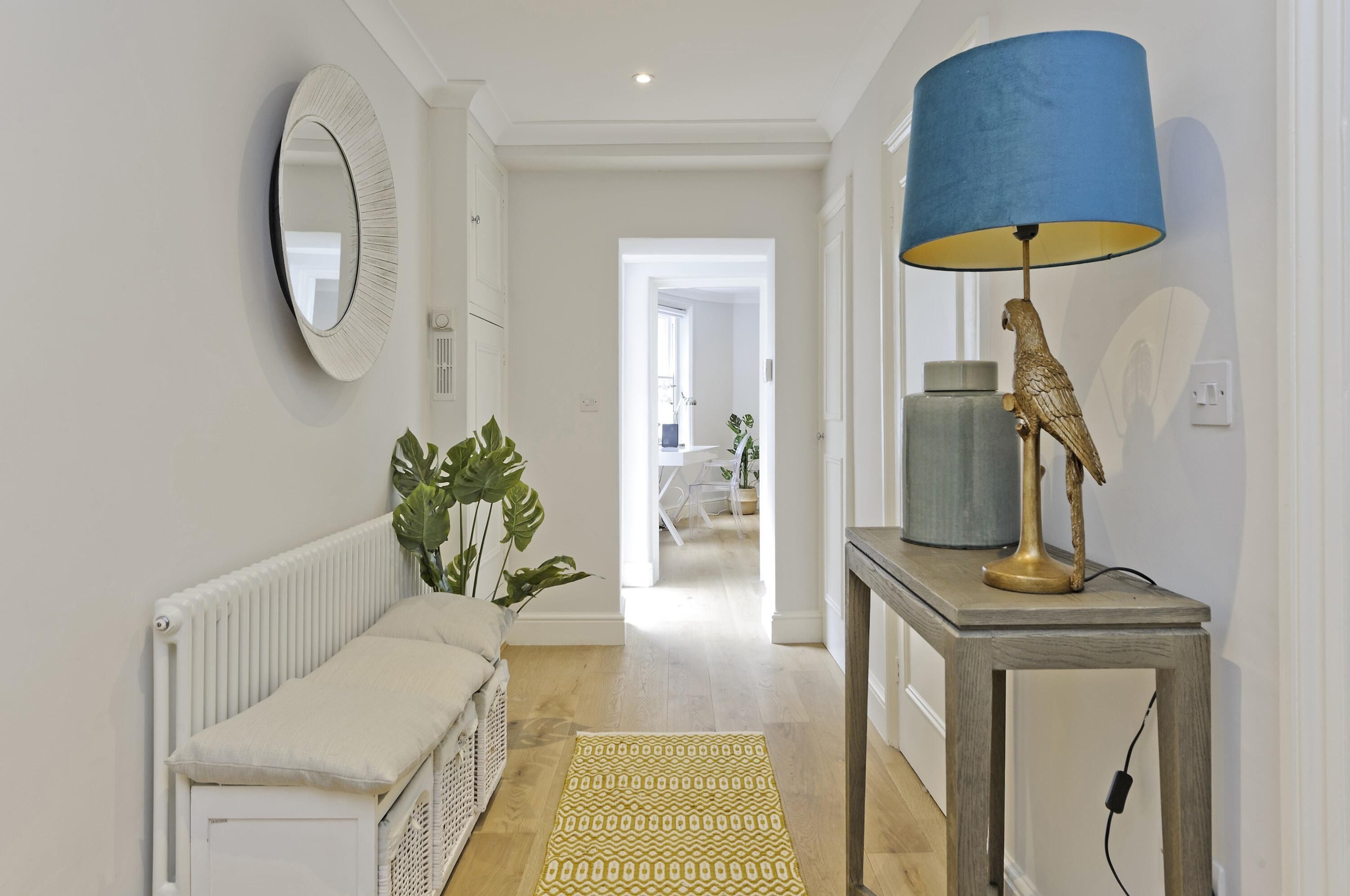 Un couloir avec un radiateur blanc, un banc beige, un tapis à motifs, une lampe bleue et une statuette d’oiseau décorative posée sur une table.