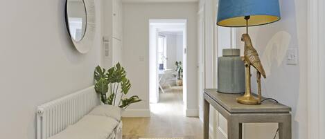 Un couloir avec un radiateur blanc, un banc beige, un tapis à motifs, une lampe bleue et une statuette d’oiseau décorative posée sur une table.