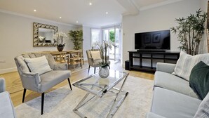 A modern living room with a glass coffee table, grey sofas, a TV, and a dining area with chairs and a table. - Huge Kensington Garden Flat (London)