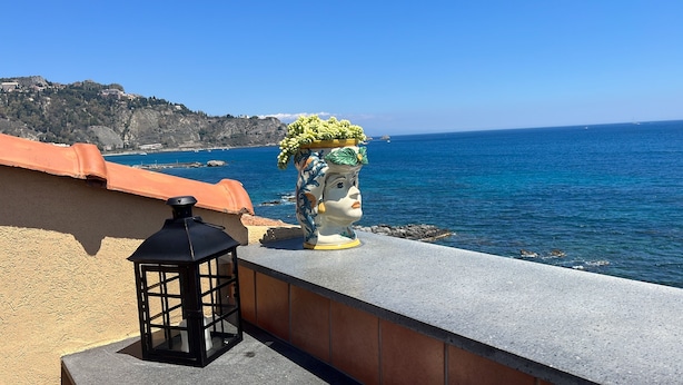 A coastal terrace with a decorative ceramic head, a black lantern, and a view of the sea and distant buildings. - Sea View Flat (Giardini Naxos)