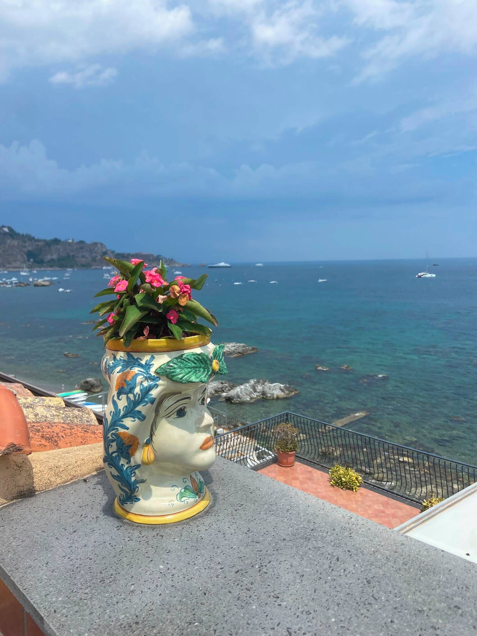 Eine Keramikvase mit einem Gesichtsmotiv, platziert auf einem Balkon mit Blick auf die Küste, wo Boote und Klippen zu sehen sind.