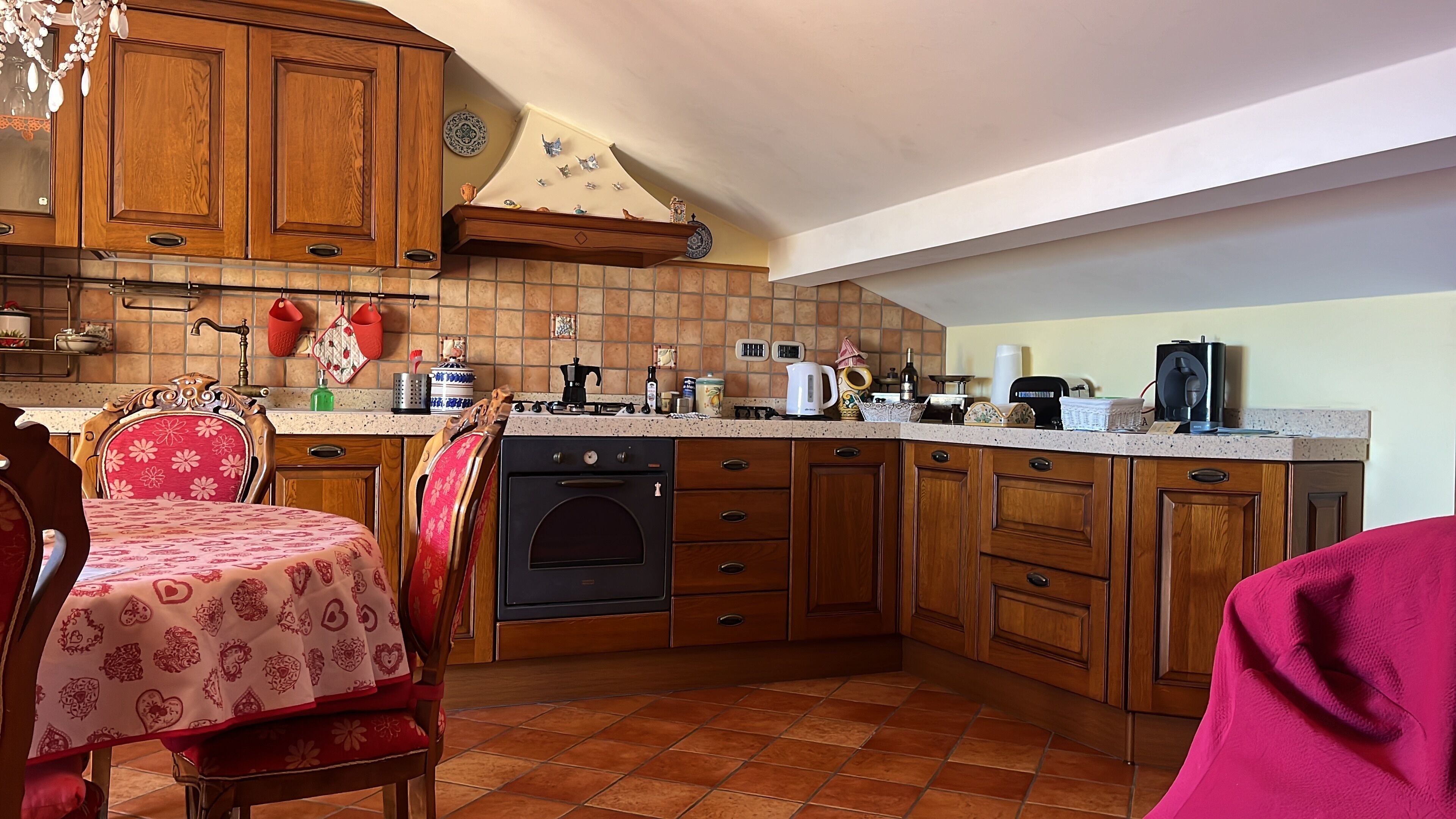 A kitchen with wooden cabinets, a built-in oven, a dining table with a floral tablecloth, and a red chair.