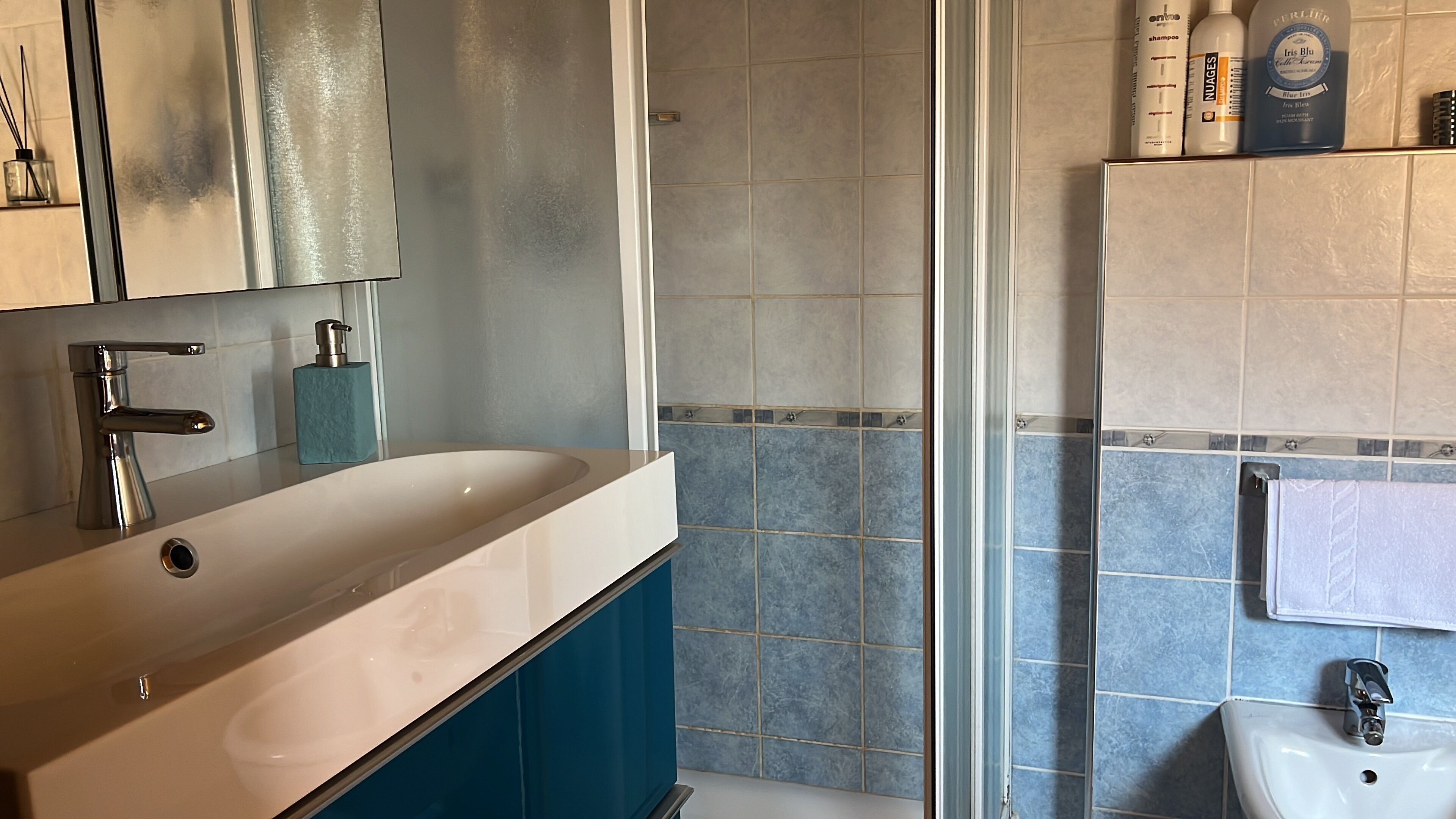 A modern bathroom with a large white sink, a blue cabinet, a glass shower enclosure, and a small white pedestal sink.