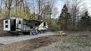 Ein Wohnmobil steht in einem bewaldeten Gebiet, in der Nähe befinden sich ein Picknicktisch und blaue Stühle.