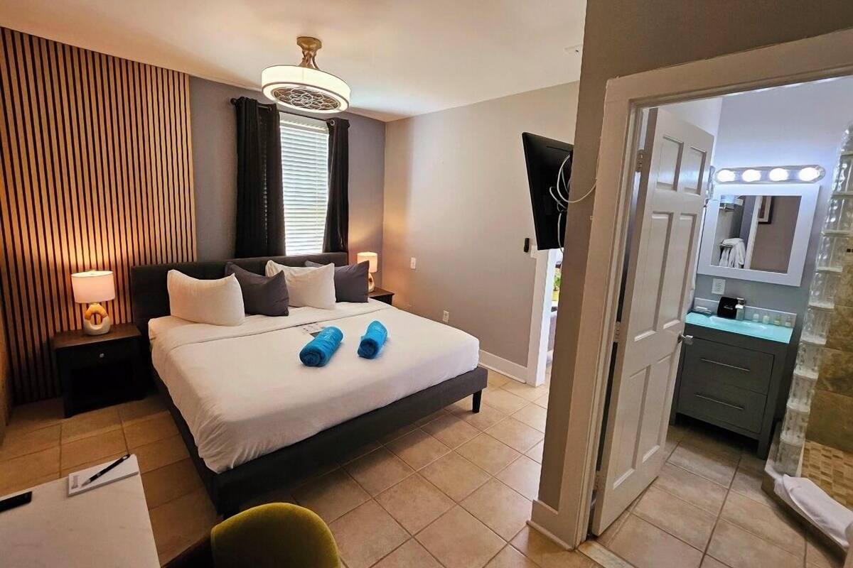 A hotel room with a bed, a nightstand, a lamp, a chair, a bathroom with a mirror and sink, and a ceiling fan.