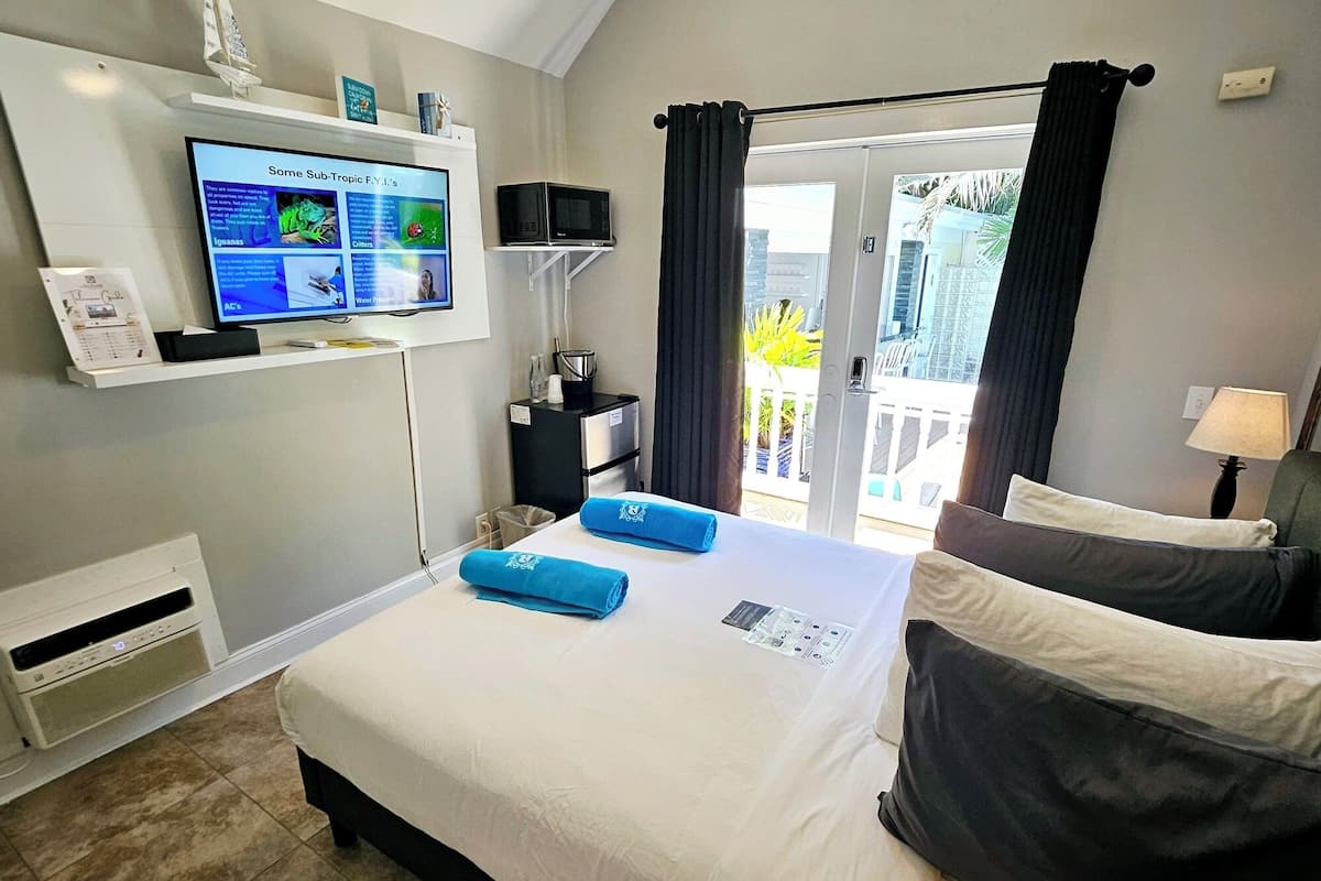 A hotel room with a bed, a television mounted on the wall, a small table with a microwave, and a view of the outside through a door.
