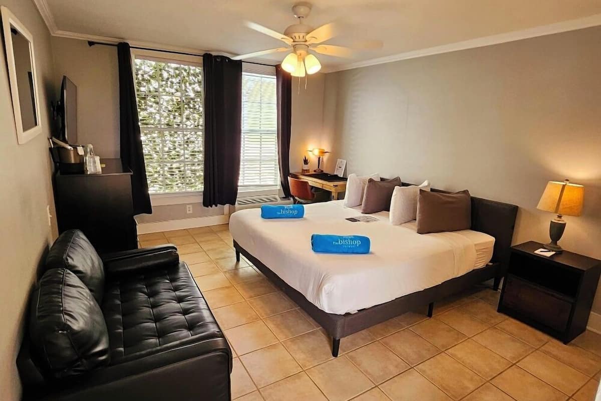 A hotel room with a bed, a desk, a chair, a black leather sofa, a nightstand, and a ceiling fan.