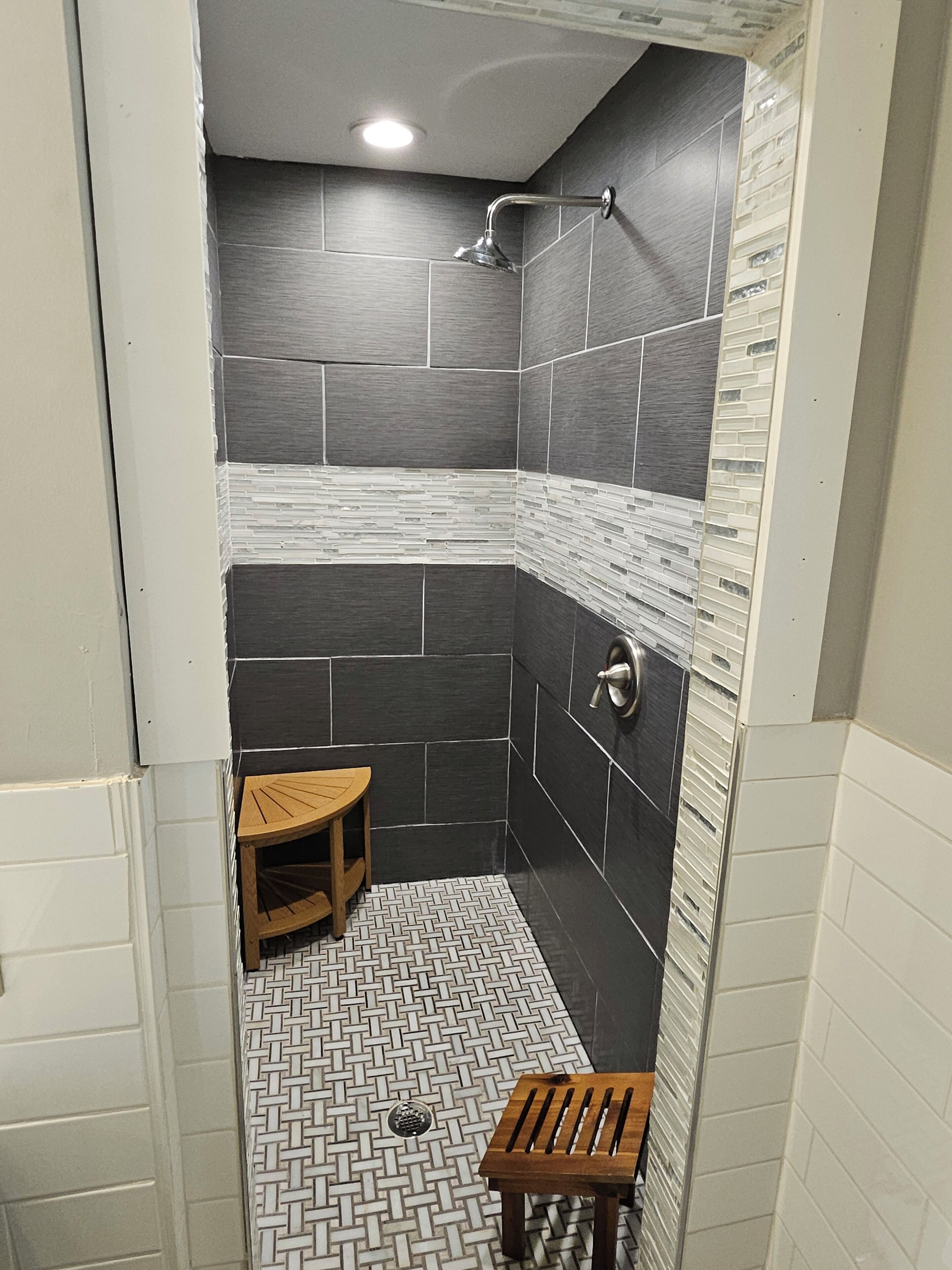 A small, modern shower with dark tiles and a wooden bench.