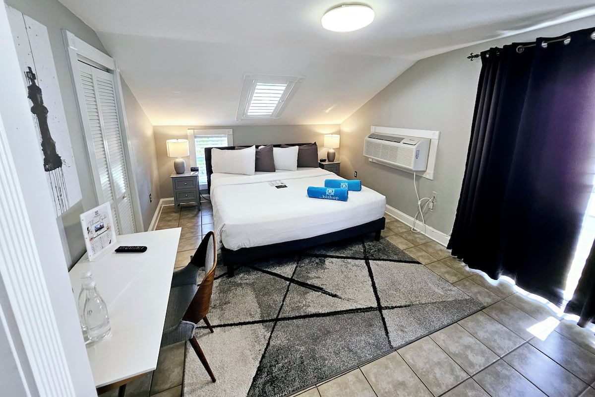 A hotel room with a large bed, a desk, a chair, a nightstand, a lamp, a window with blinds, an air conditioning unit, and a black curtain.