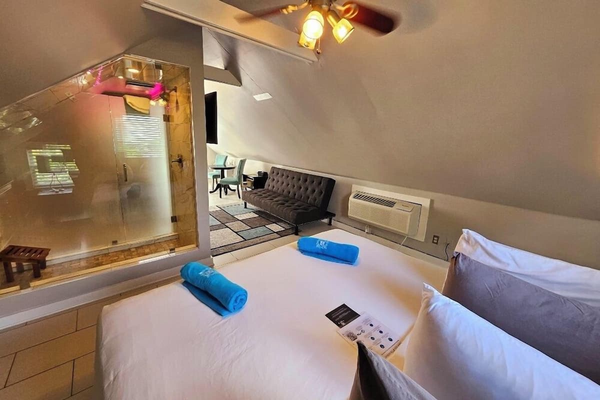 A hotel room with a bed, a ceiling fan, a window with a view of the outside, and a small table with a chair.