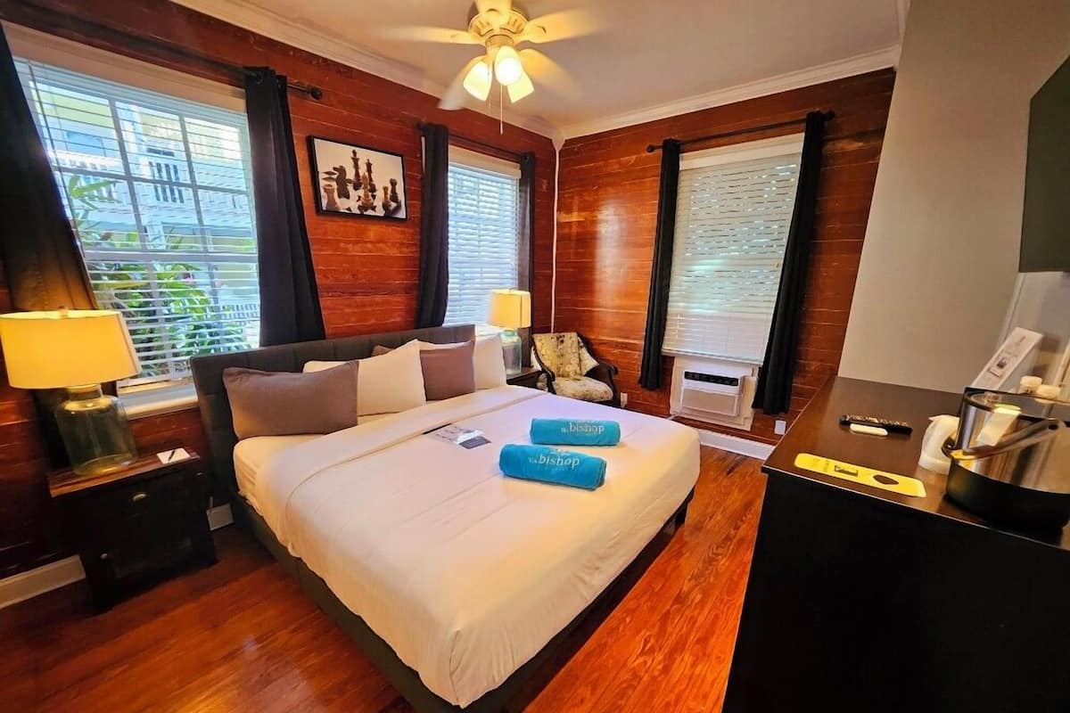 A hotel room with a large bed, wooden flooring, a ceiling fan, and a window with blinds.
