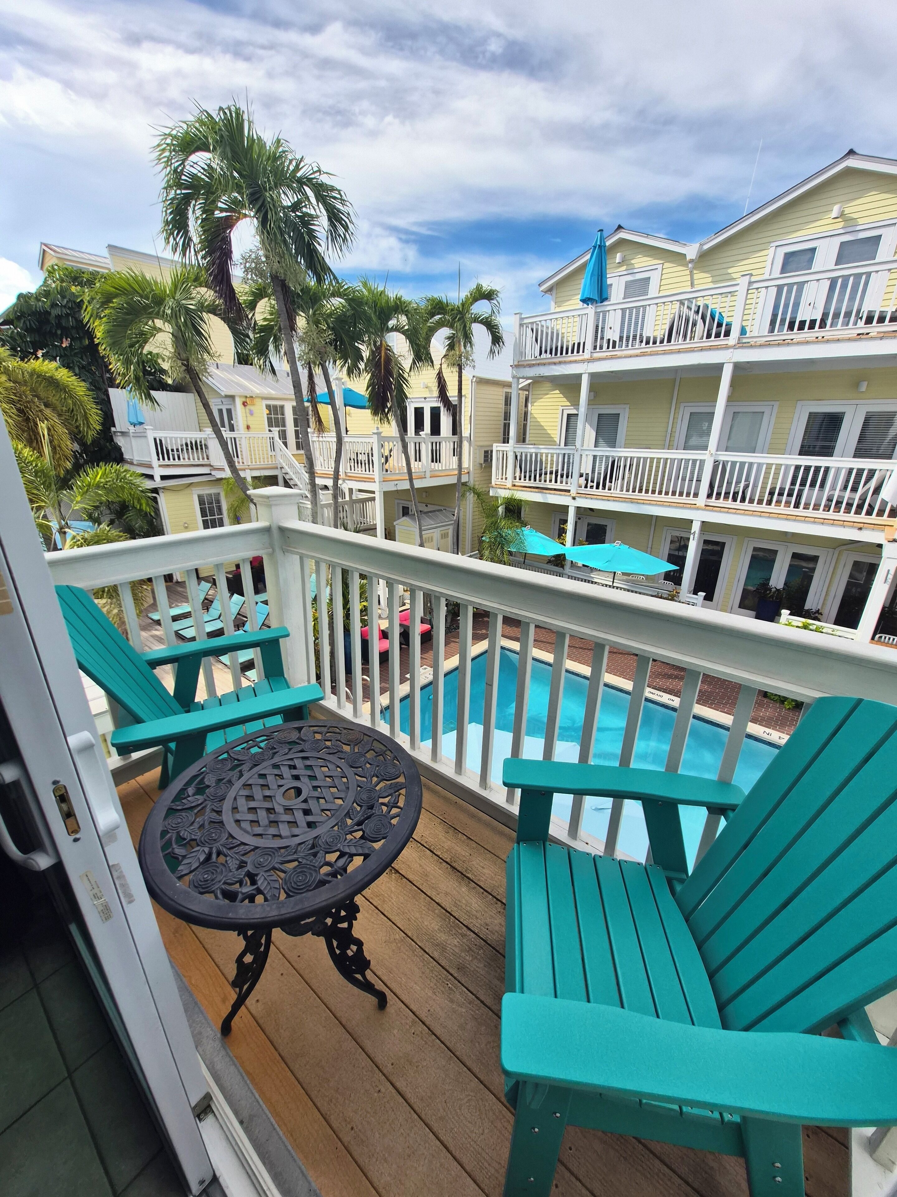 Une terrasse en bois, agrémentée de chaises turquoise et d’une table, qui offre une vue sur une piscine et un immeuble doté de balcons.
