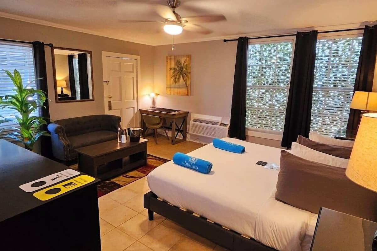 A hotel room with a bed, a desk, a chair, a sofa, a ceiling fan, and a window with blinds.