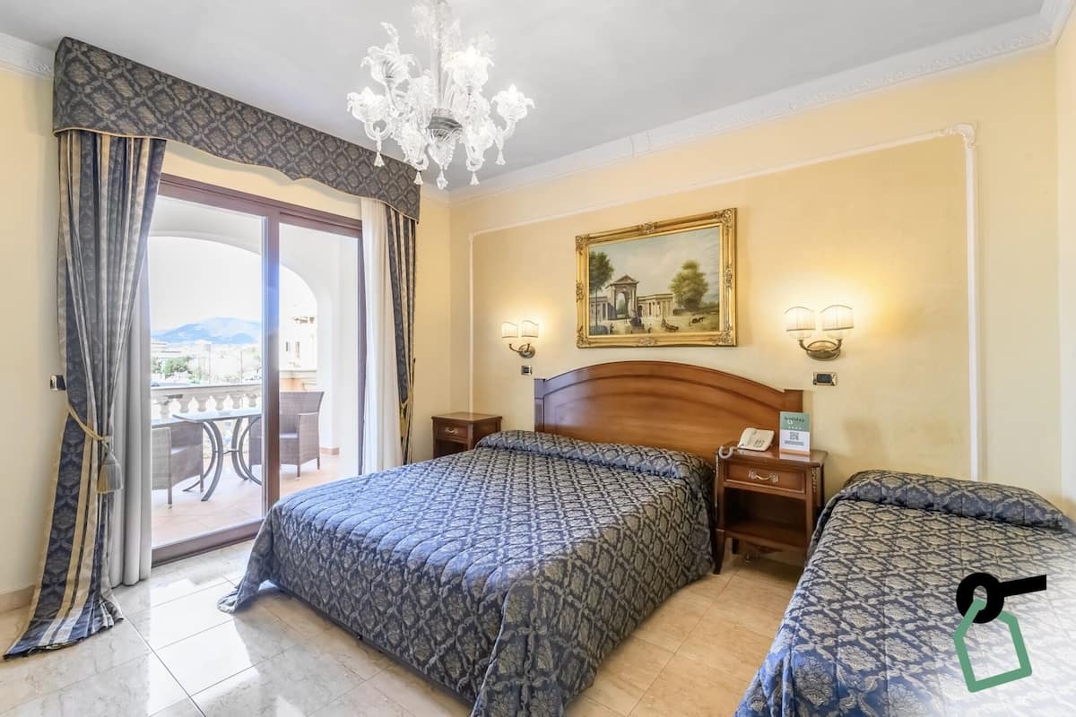 Une chambre d’hôtel avec un grand lit, un plus petit lit, un lustre, un tableau au mur et un balcon avec vue.