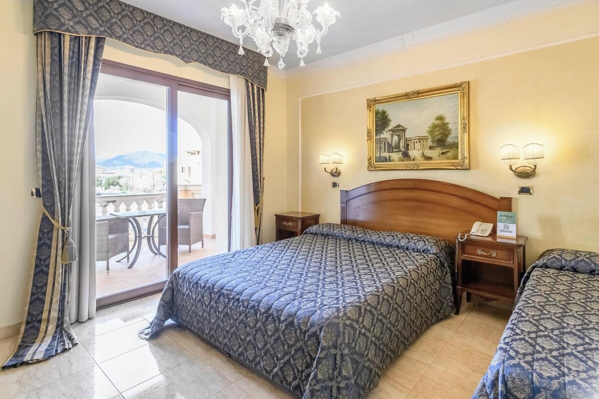 A hotel room with a large bed, a smaller bed, a chandelier, a painting on the wall, and a balcony with a view.