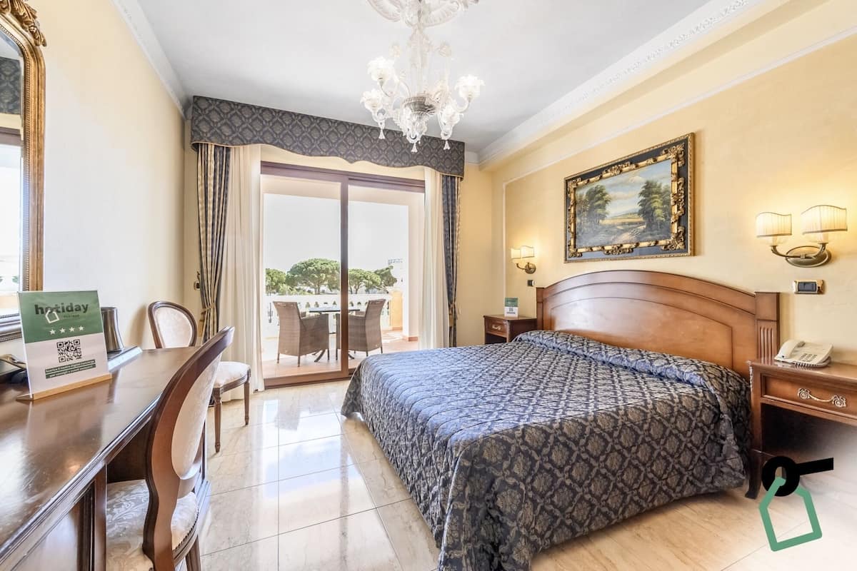 Une chambre d’hôtel avec un lit, un bureau, une chaise, un lustre, un tableau et une vue sur l’extérieur.