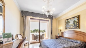 Habitación de hotel con cama, escritorio, silla, candelabro, cuadro y vistas al exterior.