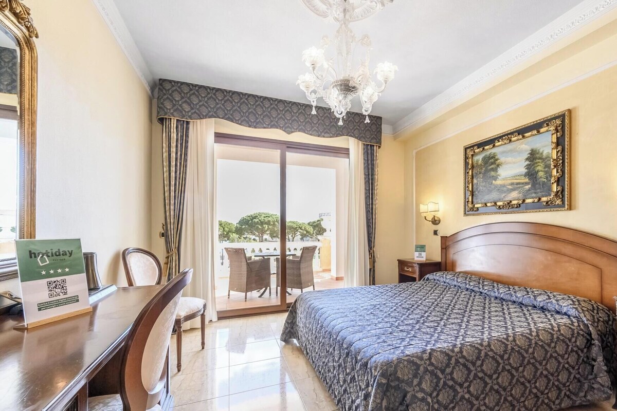 A hotel room with a bed, a desk, a chair, a chandelier, a painting, and a view of the outdoors.
