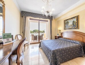 A hotel room with a bed, a desk, a chair, a chandelier, a painting, and a view of the outdoors. - Hotiday Olbia Porto (Olbia)