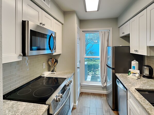 A modern kitchen with white cabinets, a built-in microwave, a stainless steel stove, and a window with curtains. - Relaxing Palms by Ryson Vacation Rentals (Galveston)