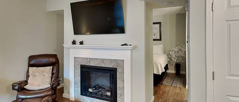 A living room with a fireplace, a leather chair, a flat-screen TV mounted above the fireplace, and a hallway leading to another room.