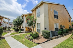 A two-story building with a beige exterior, a covered outdoor area with stairs, and a well-maintained garden with various plants and flowers. - Relaxing Palms by Ryson Vacation Rentals (Galveston)