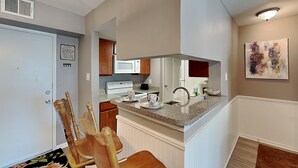 A compact kitchen with a microwave, stove, and sink. There is a wooden chair and a floral rug. - Canvas Palms by Ryson Vacation Rentals (Galveston)