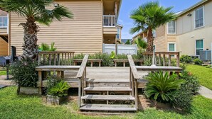 A wooden deck with steps leading to a balcony, surrounded by palm trees and a well-maintained garden. - Canvas Palms by Ryson Vacation Rentals (Galveston)