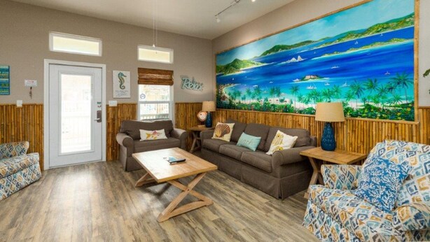 Een ruime woonkamer met een groot schilderij van een tropisch strand, een plafondventilator en een houten vloer.