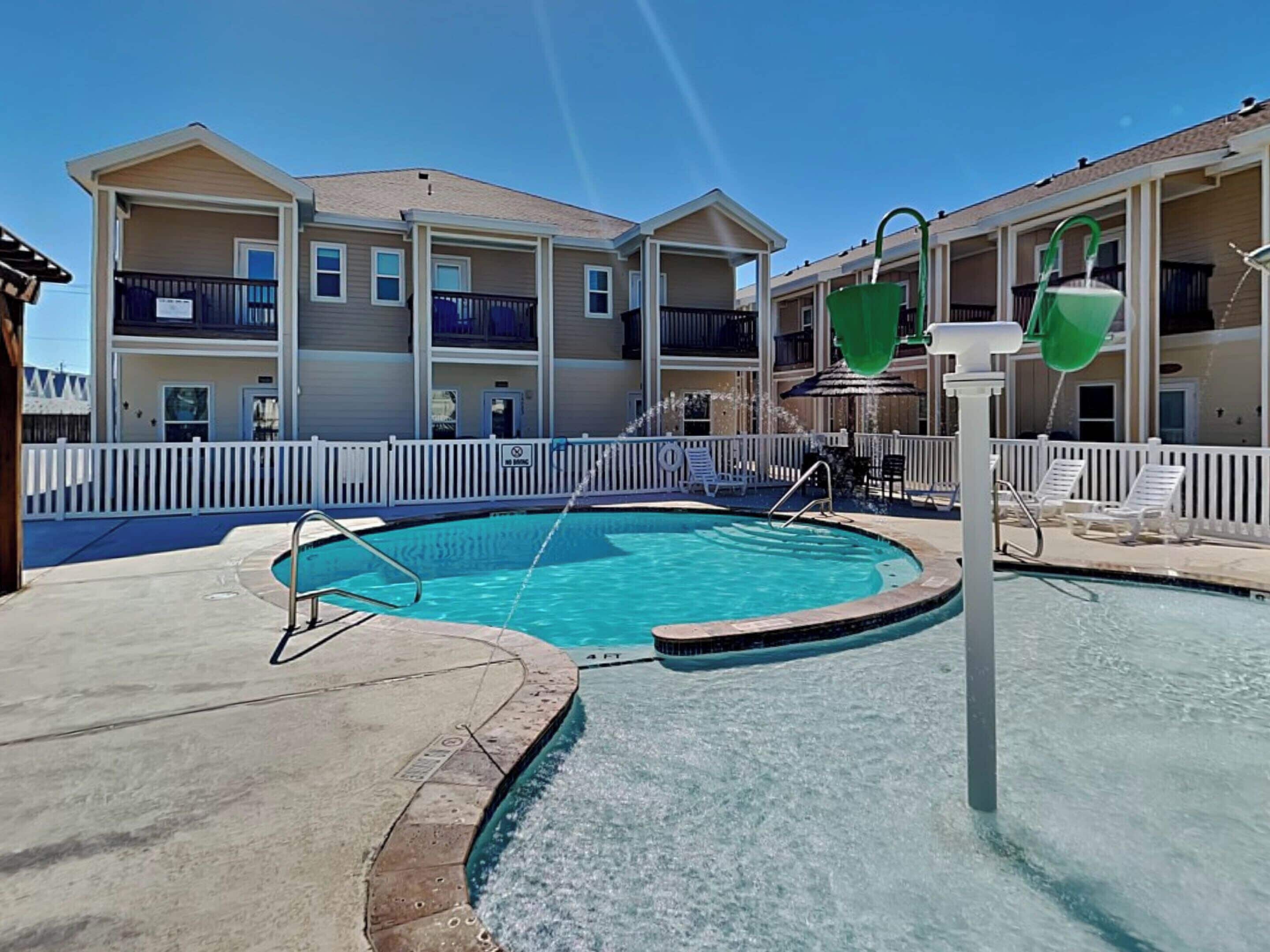 A swimming pool with a white fence and lounge chairs, surrounded by multi-story buildings with balconies.