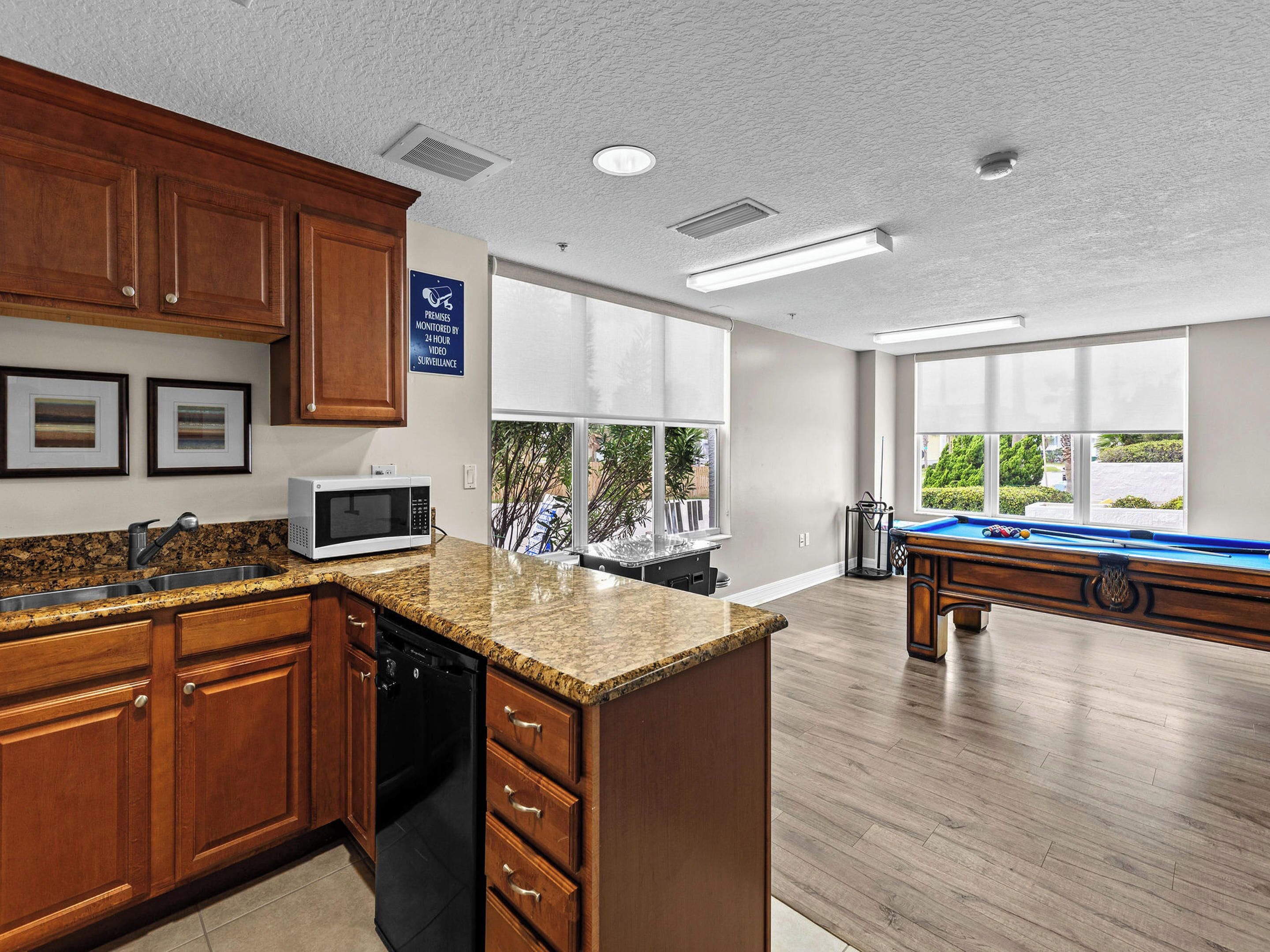 A kitchen with granite countertops, wooden cabinets, a microwave, and a dishwasher. There is a pool table in the adjacent room with large windows.