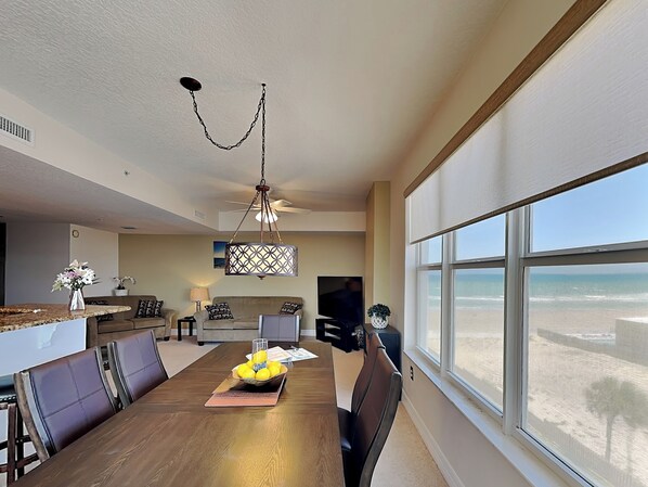 Uma sala de jantar com mesa, cadeiras, um lustre, uma TV e vista para a praia.