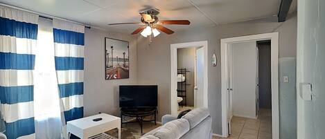 Un salon avec un canapé, un téléviseur, un ventilateur de plafond et des rideaux à rayures.