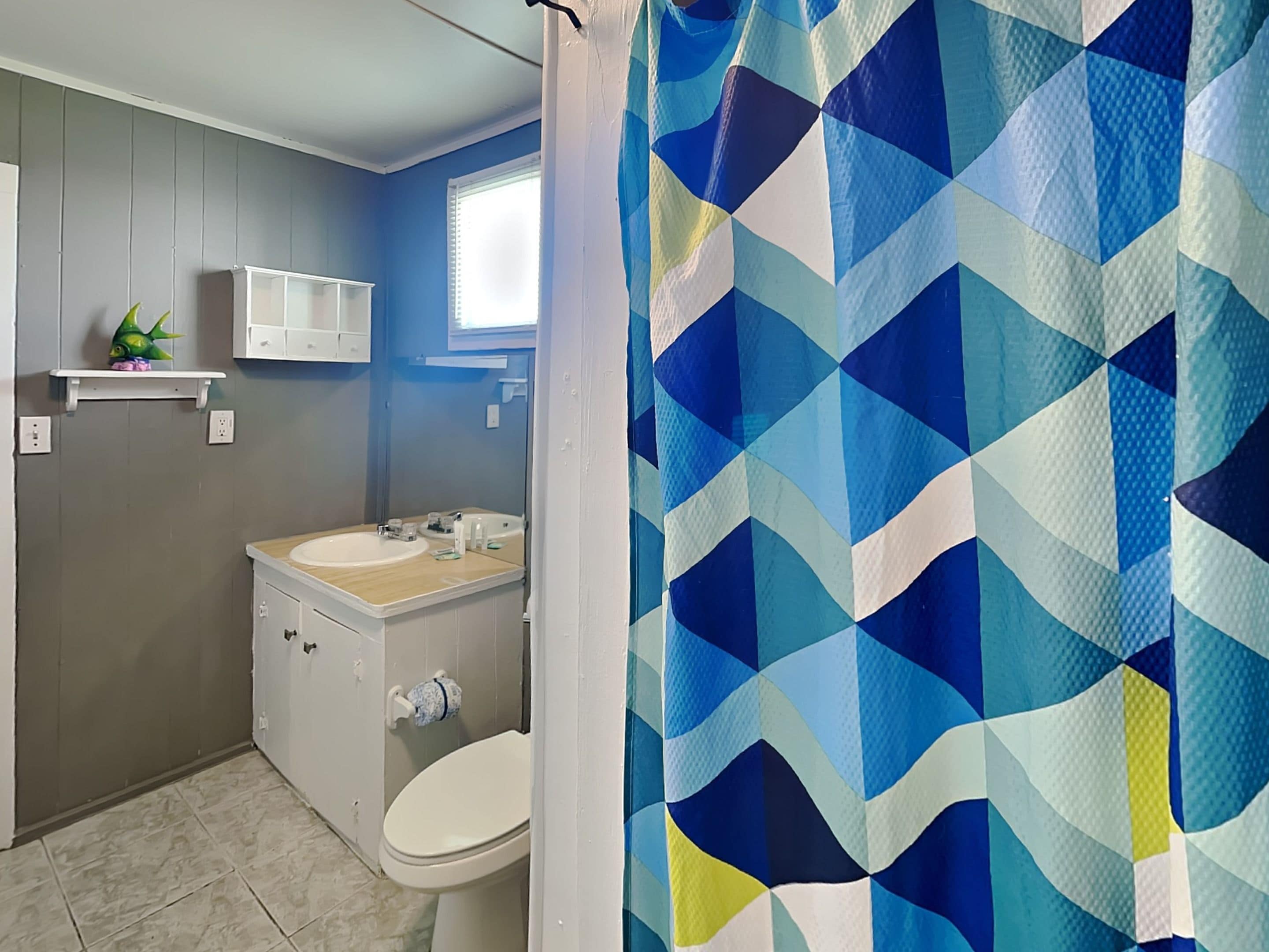 Un baño con lavamanos, inodoro y tocador blancos. Hay una ventana sobre el lavamanos y una cortina de ducha con un diseño geométrico.